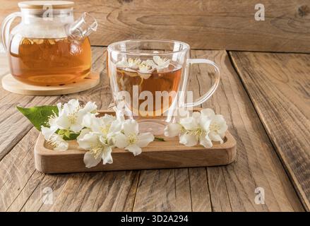 Glass transparent cup with fragrant jasmine tea on a wooden tray with the flowers of the plant. wooden rustic table with teapot. tea composition Stock Photo