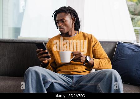 African American man in mustard sweatshirt sitting on couch checking smartphone and holding mug. Cozy, relaxation, lifestyle, modern, urban, leisure, comfort || Model released Stock Photo
