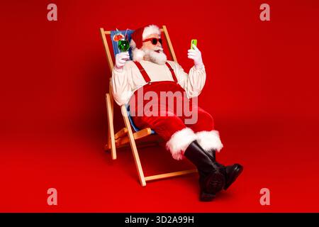 Santa relaxes in red suit on a rocking chair holding a phone and a drink in a festive Christmas scene against a bold red background Stock Photo