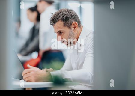 Focused professional works on laptop in a modern office with colleagues in the background Stock Photo
