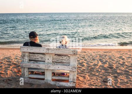 Two elderly people sit on a rustic wooden bench on a sandy beach, gazing at the calm ocean under a warm, clear sky. Stock Photo