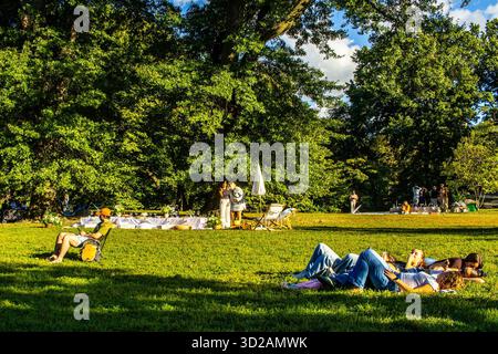 People relaxing lying down in the grass in Central Park on a sunny summer weekend afternoon as a decorated table for a picnic or party is set up Stock Photo
