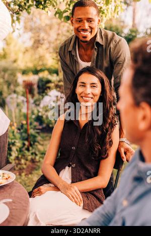 Smiling man leaning behind woman sitting on chair in back yard Stock Photo