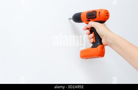Hand holding a cordless drill with orange accents, preparing to fasten a screw into a white wall, showcasing home improvement and DIY project engageme Stock Photo