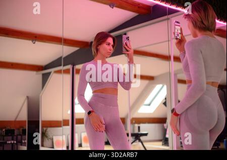 Focused woman in gray workout attire taking a selfie in a gym with mirrors. Stock Photo
