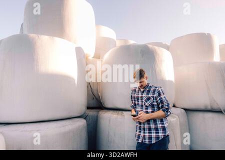 Male farmer using smart phone while leaning on hay stack in farm Stock Photo
