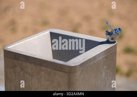 A clean, static shot of an empty public drinking fountain made of concrete and stainless steel. The scene is set outdoors on a bright day with a blurr Stock Photo