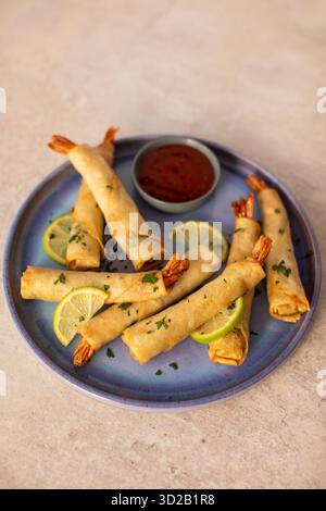 Crispy shrimp spring rolls or firecracker prawns wrapped in pastry with tails showing, garnished with fresh herbs and lime slices on a blue ceramic pl Stock Photo