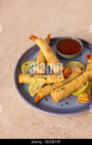 Crispy shrimp spring rolls or firecracker prawns wrapped in pastry with tails showing, garnished with fresh herbs and lime slices on a blue ceramic pl Stock Photo
