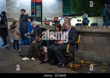 A business man sits amongst other travellers all looking at their smartphones whilst sitting on a bench on a winter's evening at Green Park, London UK. Stock Photo