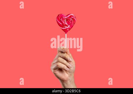 A Caucasian man's hand holds a shiny red and pink heart-shaped lollipop candy, isolated on a vibrant solid pink-red background for Valentine's Day. Stock Photo