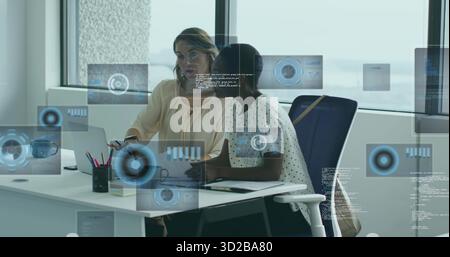 Analyzing data, two women in blouses collaborating at office desk, with laptop, floating graphics Stock Photo