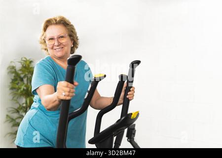 A woman is smiling while holding the handlebars of a stationary bike. Wearing a blue T-shirt Stock Photo