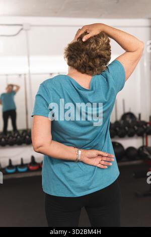 A woman is in a gym, wearing a blue shirt and black pants. She is bending over and touching her head with her hand. Concept of determination and focus Stock Photo