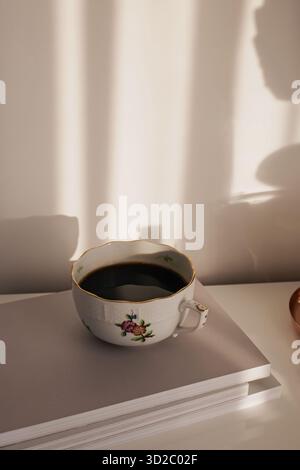 A delicate porcelain tea cup with floral patterns filled with coffee rests on a stack of books. Stock Photo
