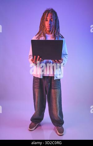 Teenager with dreadlocks standing and concentrating on a laptop screen with neon light reflections Stock Photo