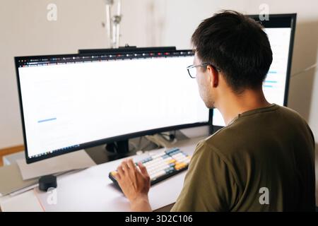 View from shoulder of programmer in glasses concentrating on complex task, typing code on mechanical keyboard in home office, demonstrating modern Stock Photo
