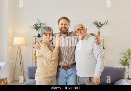 Millennial man hugging his older parents standing together in living room Stock Photo