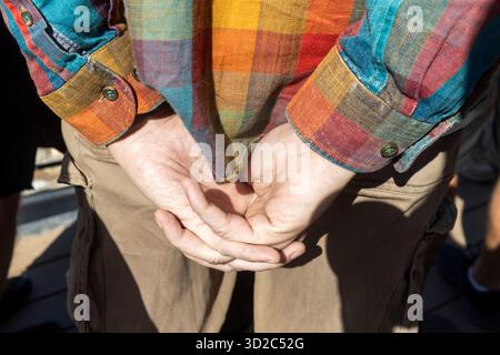 A close-up view of a person's hands clasped behind their back, wearing a brightly colored, checkered flannel shirt with dominant red, orange, yellow, Stock Photo