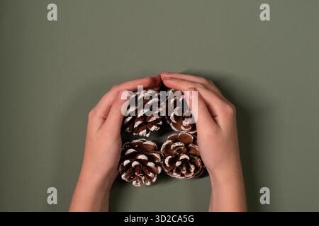 Child's hands holding pine tree cones on a green olive background with copy space. Christmas minimalistic aesthetic template. Flat lay, top view Stock Photo