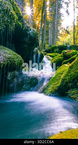 Mossy rocks and waterfall illuminated by sunlight streaming through dense autumn forest Stock Photo