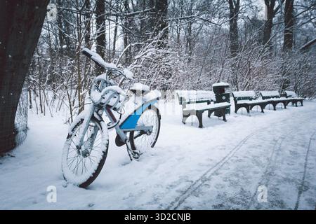 Snow-covered park path in winter with benches and a bicycle parked on the side. Everything around: the path, trees, and benches - is blanketed in snow Stock Photo