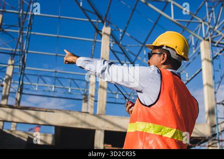 Male Construction worker or Engineer or Foreman stands at a building site  using a walkie-talkie radio to coordinate and consulting with the team Stock Photo