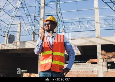Male Construction worker or Engineer or Foreman stands at a building site  using a walkie-talkie radio to coordinate and consulting with the team Stock Photo