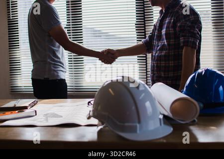 Two construction professionals shaking hands over blueprints and safety equipment signifying a successful project agreement and partnership Stock Photo