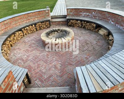 A cozy and rustic outdoor fire pit area with a circular design. The central brick fire pit is surrounded by curved wooden benches. Stock Photo