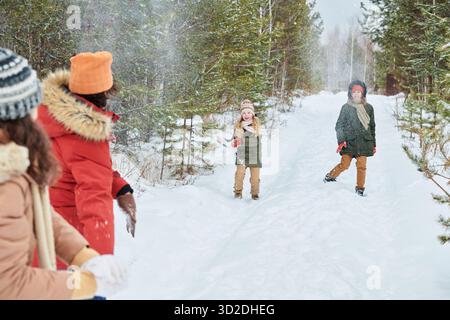 Two Caucasian children playing in snow on forest path while Caucasian parents watching and smiling, winter trees surrounding group, snow falling gently around scene Stock Photo
