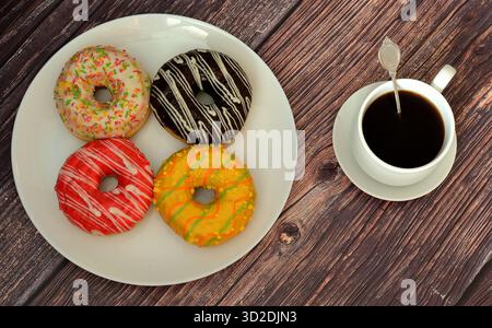 Four multi-colored glazed doughnuts with toppings on a round plate and a cup of hot black coffee on a wooden table. Top view, flat lay. Stock Photo