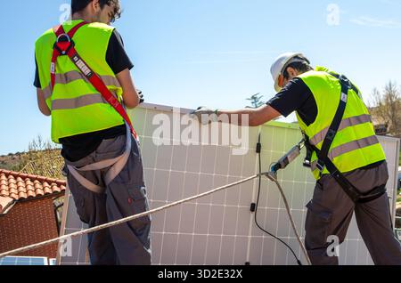 Two workers wearing safety vests and harnesses secure solar panels onto a rooftop under clear blue sky in a residential neighborhood. The sun is shini Stock Photo