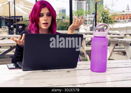 Woman with vibrant magenta hair showing anger while working remotely using a laptop at an outdoor co-working space table Stock Photo