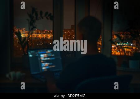 The blurred code is on a laptop on a wooden table in front of the window in the dark with a view of the lights of the night city. Focus on the city vi Stock Photo
