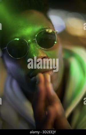 Hopeful greenlit African man with folded hands wearing sunglasses at night as shade of anonymity, praying, approved immigration, anticipation, expecta Stock Photo