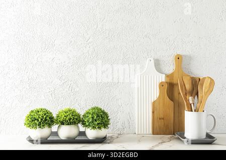 Cutting boards and wooden spoons in a white mug on a marble white countertop against a gray cement wall with green houseplants Stock Photo
