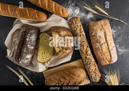 An assortment of baked bread on a black background with scattered flour and ears of corn. delicious homemade bread Stock Photo