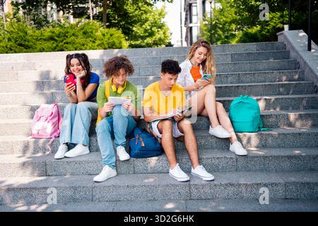 Young group of friends sitting on steps outdoors, enjoying social media, studying, and bonding during the sunny day Stock Photo