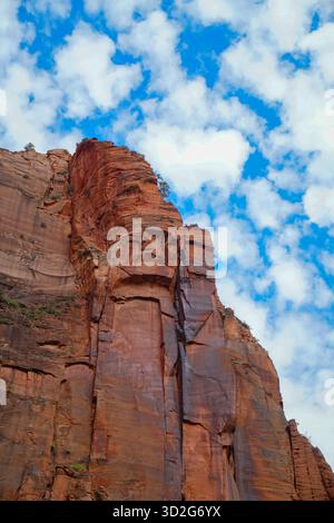Sunlit red sandstone cliffs rise sharply against a cloud-filled sky in Zion National Park, emphasizing dramatic textures, layered rock patterns, and b Stock Photo