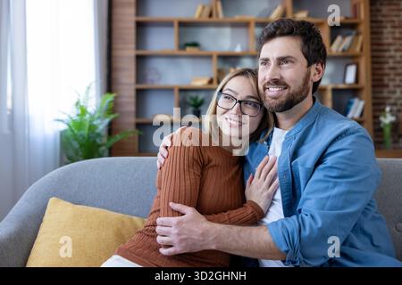 Loving couple sitting on a cozy sofa, hugging and smiling, looking away with happy expressions, embodying comfort, strong relationship, and a shared future inside a modern living room Stock Photo