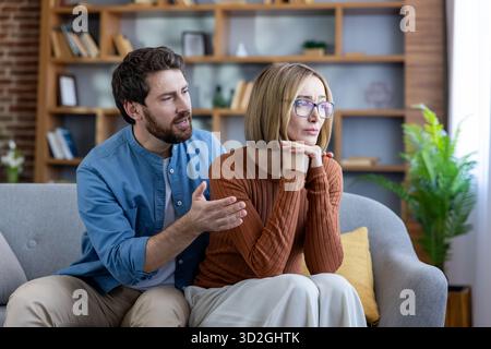 Man reaches out to upset partner on sofa as she turns away, tense couple after disagreement in living room, emotional moment of apology, support and attempted reconciliation Stock Photo