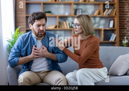 Man experiencing acute chest pain and holding his chest with both hands while sitting on a couch, with a worried woman supporting him by placing her hand on his shoulder Stock Photo