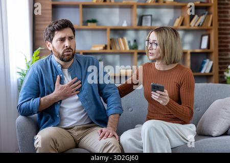 Couple sitting on a couch at home, man feeling chest pain and breathing difficulty while woman holds a smartphone, both reacting to shocking, stressful information or health problems Stock Photo