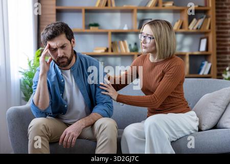Woman supporting and comforting a man experiencing stress and headache, sitting on a sofa in a home setting, symbolizing empathy and relationship challenges Stock Photo