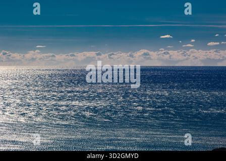 A wide seascape with a clear horizon line. The deep blue surface of the water reflects bright sun glare. A horizontal strip of white clouds stretches Stock Photo