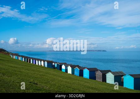 A row of colourful wooden beach huts in Tankerton, Whitstable at the bottom of a grass verge along a long beach. The sea and sky are blue. Isle of She Stock Photo