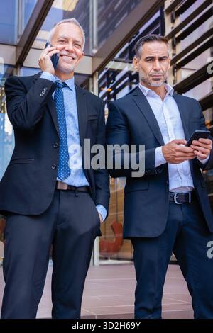 Two businessmen in suits using smartphones, talking outside modern office. Stock Photo
