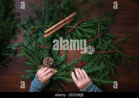 Hands of a mature woman arranging evergreen branches with decorations for an Advent wreath, preparation for Christmas on a brown wooden table, top vie Stock Photo