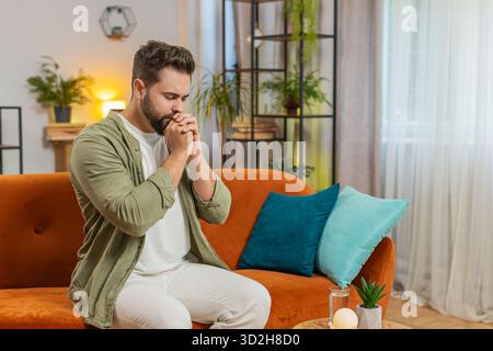 Tensed young man sitting on sofa at home folding palms together pray ask for help, thinks feel stressed looks desperate by life problems. Caucasian guy praying asking for help sitting on sofa at home. Stock Photo
